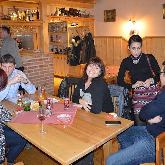 Eine Gruppe von Menschen sitzt an einem Tisch in einem Restaurant. Eine Frau steht und spricht mit einem Mann. Auf dem Tisch stehen ein Weinglas, eine Flasche und ein Handy. Hinter ihnen steht ein Mann hinter der Theke.