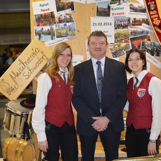 Ein Mann in einem Anzug steht zwischen zwei Frauen in traditionellen Kostümen. Sie stehen vor einer Wand mit verschiedenen Fotos und einem Schild, das 'Musveren Schwodorf' besagt. Im Hintergrund sind Musikinstrumente zu sehen.