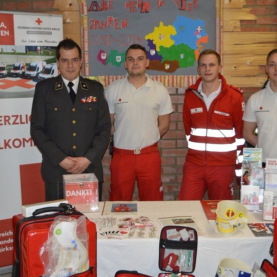 Vier Männer in Uniformen stehen hinter einem Tisch mit verschiedenen medizinischen Utensilien und Informationsmaterialien. Ein Mann trägt eine Militäruniform, während die anderen rote und weiße Uniformen tragen. Ein Banner mit dem Text 'Herzlich Willkommen' ist im Hintergrund sichtbar.