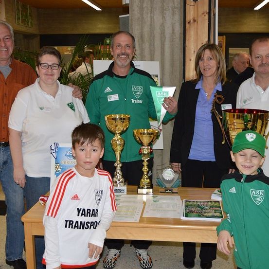 Eine Gruppe von Menschen steht um einen Tisch mit Trophäen und Auszeichnungen. Zwei Jungen in Sporttrikots stehen vorne. Ein Mann in einem grünen Trikot hält ein Zertifikat. Eine Frau in einem schwarzen Blazer lächelt.