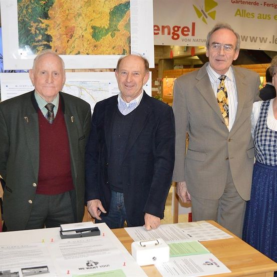 Vier Personen stehen vor einem Tisch mit Papieren und einem Schild, das Engel sagt. Der Mann auf der linken Seite trägt eine grüne Jacke und der Mann auf der rechten Seite trägt ein blaues Kleid.