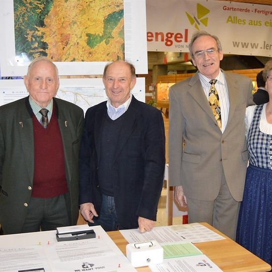 Vier Personen, zwei Männer und zwei Frauen, stehen zusammen vor einem Tisch mit Papieren und einem Schild, das 'Engel' steht. Der Mann links trägt eine grüne Jacke, der Mann in der Mitte trägt einen dunklen Anzug, der Mann rechts trägt einen hellen Anzug und eine Krawatte, und die Frau rechts trägt ein blaues Kleid. Hinter ihnen ist eine Wand mit einer Karte und einem Banner.