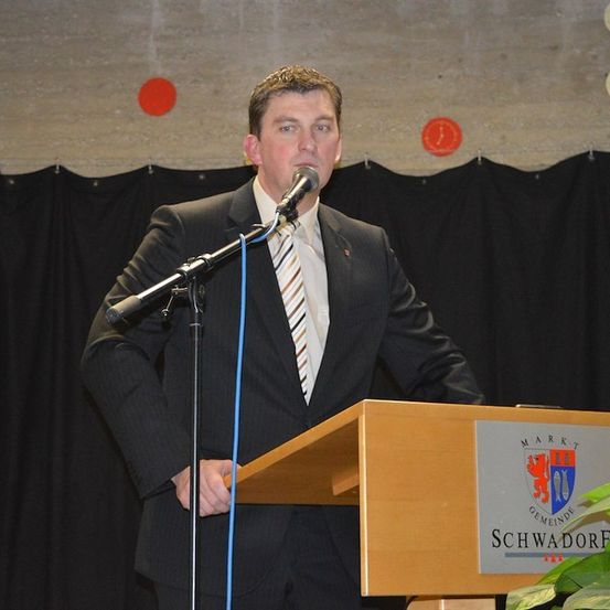 Ein Mann in einem Anzug steht an einem Podium mit einem Mikrofon und spricht hinein. Hinter ihm befindet sich ein Hintergrund mit einem Logo und Wappen.