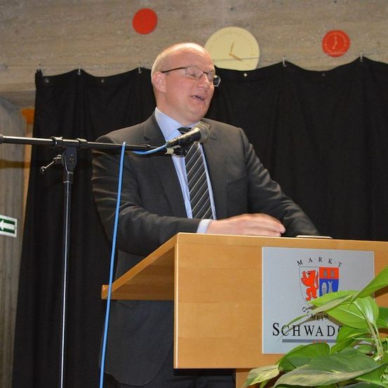 Ein Mann in Anzug und Krawatte spricht an einem Rednerpult mit Mikrofon vor einer Kulisse mit Uhr und roten Kreisen.