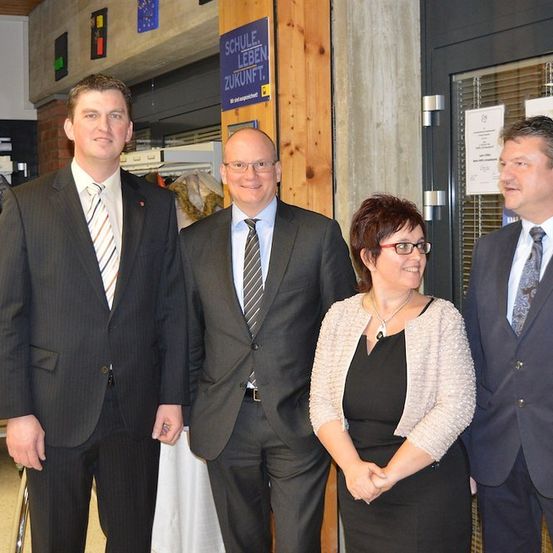 Vier Personen in formeller Kleidung stehen gemeinsam vor einem Gebäude und lächeln für ein Foto. Dahinter steht ein Schild mit der Aufschrift 'SCHULE LEBEN ZUKUNFT'.