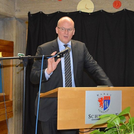 Ein Mann in Anzug und Krawatte spricht in ein Mikrofon an einem Podium mit einem Schild, das 'Markt Gemeinde' steht. Dahinter ist ein schwarzer Vorhang und eine Wand mit einer Uhr.