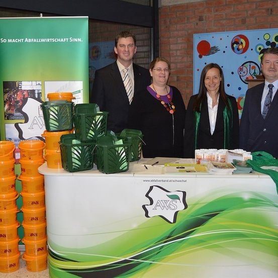 Vier Personen stehen hinter einem Tisch mit Werbematerialien, darunter grüne Körbe und orangefarbene Eimer, vor einer Ziegelwand.