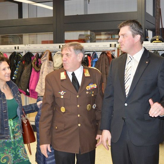 Drei Personen, zwei Männer in Anzügen und eine Frau, stehen vor einem Kleiderständer mit verschiedenen Mänteln. Der Mann in der Mitte trägt eine Militäruniform.