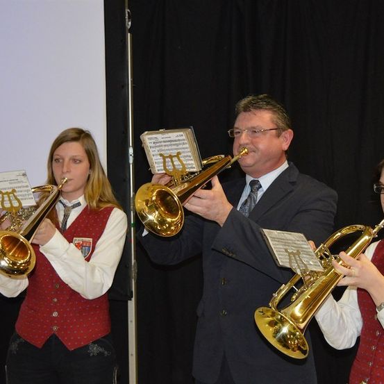 Drei Musiker spielen auf der Bühne Posaunen. Der Mann in der Mitte trägt eine Brille und einen Anzug. Die beiden Frauen tragen rote Westen.