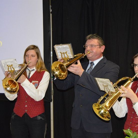 Drei Musiker, zwei Frauen und ein Mann, spielen Trompeten auf der Bühne. Der Mann steht in der Mitte, und die Frauen sind auf beiden Seiten. Sie tragen alle formelle Kleidung.