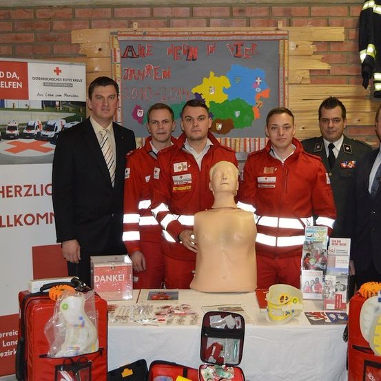 Eine Gruppe von Männern in roten Uniformen steht hinter einem Tisch mit einem Mannequinkopf, medizinischen Kits und anderen Gegenständen. Hinter ihnen befindet sich eine Ziegelwand mit einem Schild, das 'Herzlich Willkommen' besagt.