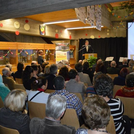 Ein Mann spricht an einem Podium vor einer großen Leinwand, während eine Menschenmenge aufmerksam zuhört in einem Konferenzsaal.