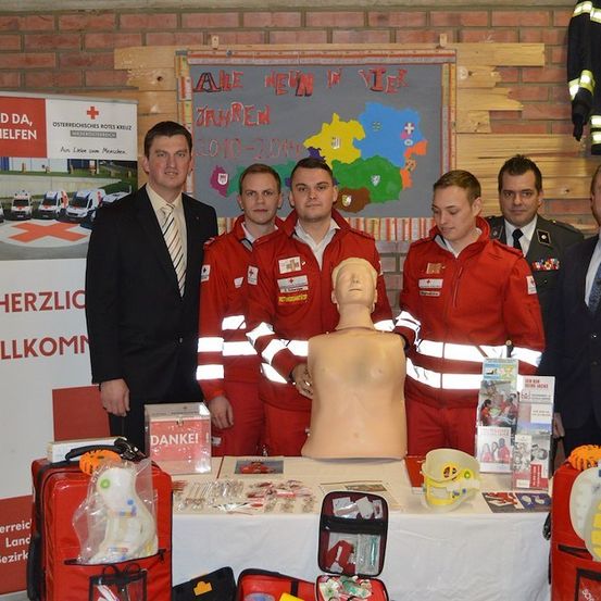 Eine Gruppe von Personen in roten Uniformen steht hinter einem Tisch mit einem Mannequin, medizinischen Kits und einer Karte. Der Schauplatz scheint eine Veranstaltung oder Ausstellung zu sein.