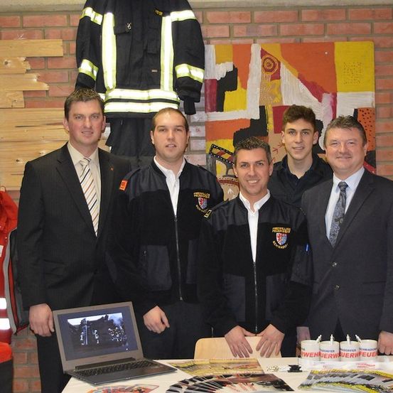 Fünf Männer in Anzügen und Jacken stehen hinter einem Tisch mit Laptop und Tassen. Ein Mann trägt eine Feuerwehrjacke. Eine Ziegelwand und ein farbenfrohes Gemälde im Hintergrund.