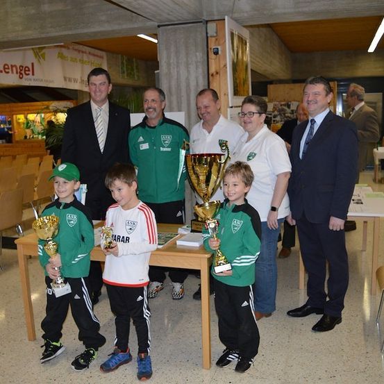 Eine Gruppe von Menschen, darunter junge Jungen, die Pokale halten, steht in einem Raum mit einem Banner, das 'Lengel' besagt. Die Jungen sind in Sportuniformen gekleidet und scheinen einen Sieg zu feiern. Dahinter stehen Erwachsene, einige mit Brillen und Krawatten, die ihre Arme umeinander geschlungen haben.