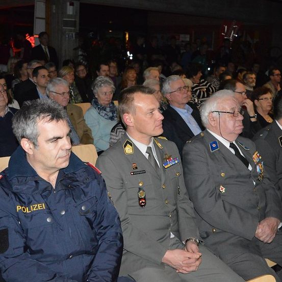 Eine Gruppe von Männern in Militäruniformen und Zivilkleidung sitzt in einer Reihe in einem schwach beleuchteten Raum, mit vielen Menschen im Hintergrund.