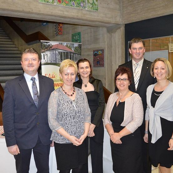 Eine Gruppe von sechs Menschen in formeller Kleidung steht lächelnd für ein Foto in einem Raum. Hinter ihnen befindet sich eine Treppe und eine Wand mit Kunstwerken.
