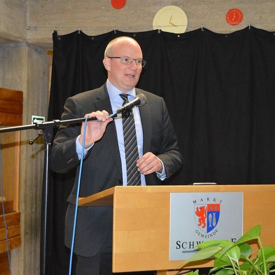 Ein Mann in Anzug und Krawatte spricht in ein Mikrofon an einem Podium mit einem Schild, das 'Marktgemeinde Schweinfurt' steht. Dahinter ist ein schwarzer Vorhang und eine Wand mit einer Uhr.