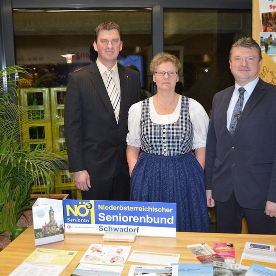 Drei Personen stehen hinter einem Tisch mit Broschüren. Ein blaues Schild mit 'Niederosterreichischer Seniorenbund Schwadorf'. Die Frau trägt ein traditionelles Kleid und eine Brille.