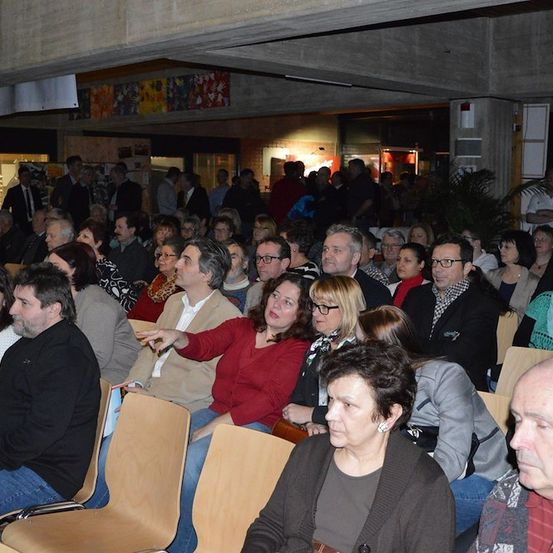 Eine große Gruppe von Menschen sitzt in einem Auditorium, einige mit Brillen und Jacken, wobei eine Frau in einem roten Shirt auf etwas vor ihr zeigt.