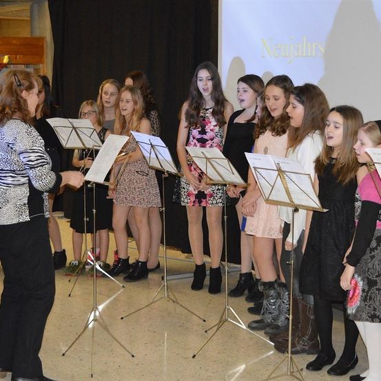 Ein Chor junger Mädchen, angeführt von einer Frau, tritt in einem Raum auf, in dem eine Projektionsleinwand 'Neujahrs' zeigt. Sie stehen vor Notenständern und sind in verschiedenen Outfits gekleidet.