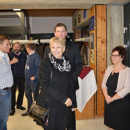 Eine Gruppe von Menschen steht in einem Raum, lächelt und unterhält sich. Eine Frau in einem schwarzen Mantel hält eine schwarze Handtasche. Ein Mann in einem Anzug steht hinter ihr. Ein Tisch mit einer weißen Tischdecke ist rechts.