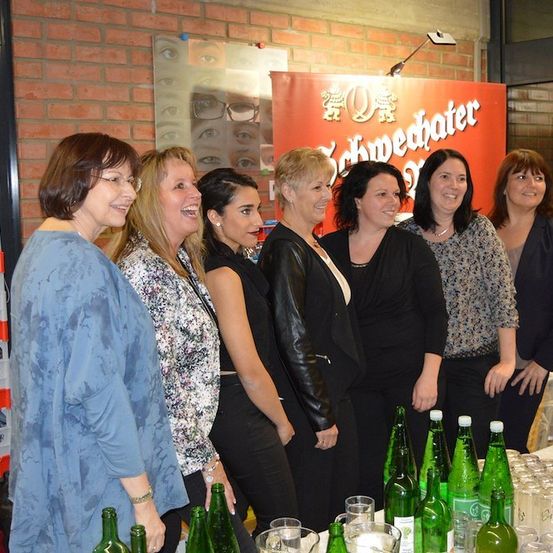 Eine Gruppe von Frauen steht hinter einem Tisch mit grünen Flaschen und Gläsern, lächelt und posiert für ein Foto vor einer Ziegelwand mit einem Schild.