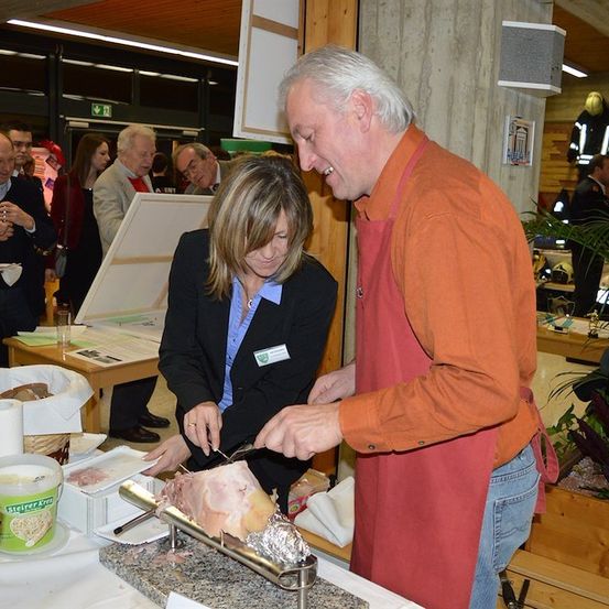 Ein Mann und eine Frau schneiden ein großes Stück Fleisch bei einer Veranstaltung, umgeben von anderen Leuten.