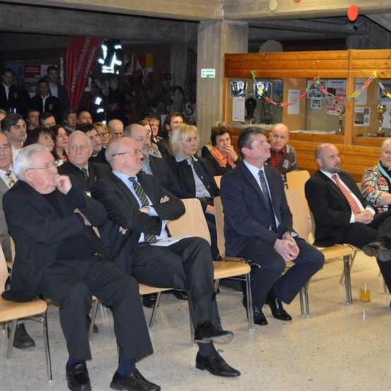 Eine Gruppe formell gekleideter Personen sitzt in Stühlen in einem Saal, möglicherweise bei einer Veranstaltung. Einige unterhalten sich, während andere sich umsehen. Ein Banner und Trophäen sind an der Wand hinter ihnen angebracht.
