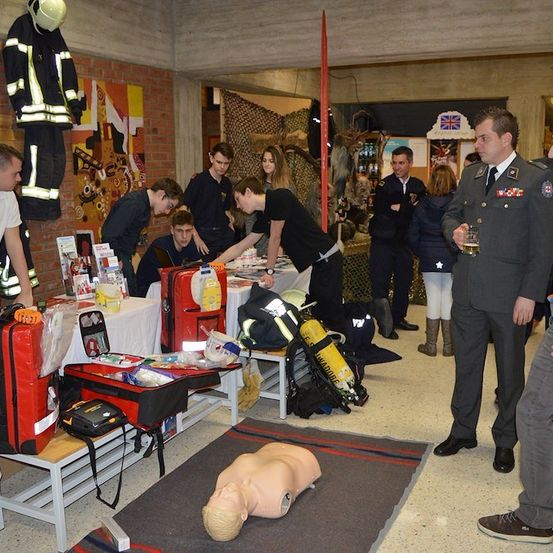 Eine Gruppe von Menschen versammelt sich um einen Tisch mit verschiedenen Notfallausrüstungen. Ein Mannequin liegt auf dem Boden, und eine Feuerwehruniform hängt an der Wand.