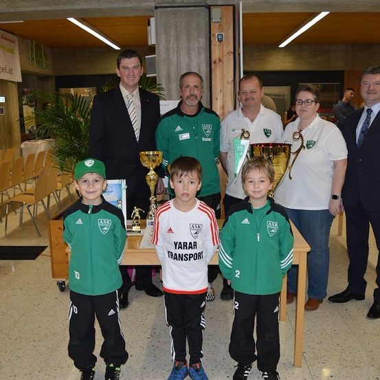 Eine Gruppe von Menschen, darunter Jungen und Erwachsene, stehen zusammen und posieren für ein Foto. Die Jungen tragen grüne und weiße Trikots mit Nummern darauf. Sie halten Trophäen in den Händen, und hinter ihnen stehen Erwachsene. Es gibt einen Holztisch mit Trophäen darauf. Links stehen Stühle und eine Topfpflanze. An der Wand hängt ein Banner.