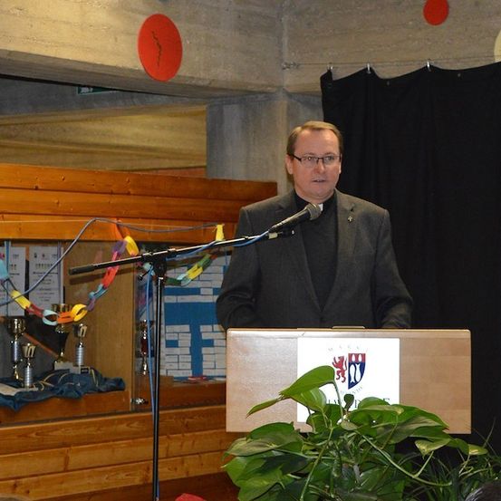 Ein Priester steht an einem Rednerpult mit Mikrofon und hält eine Rede in einem Raum mit Holzwänden und einem schwarzen Vorhang. Eine Pflanze und Trophäen sind hinter ihm.