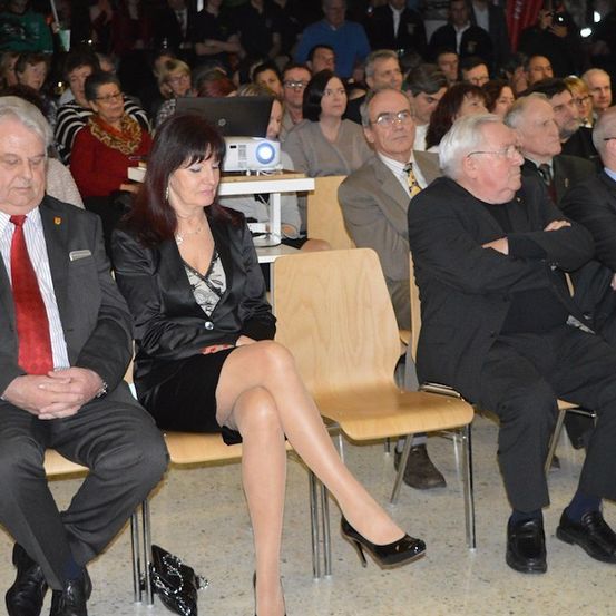 Eine Gruppe von Menschen in formeller Kleidung sitzt in Stühlen in einem Konferenzsaal, mit einem Projektor auf einem Tisch im Hintergrund.
