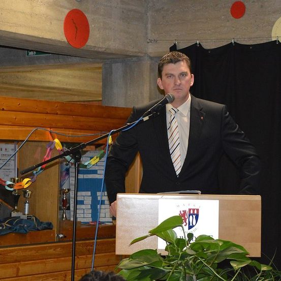 Ein Mann in einem Anzug steht an einem Podium mit einem Mikrofon, möglicherweise hält er eine Rede. Hinter ihm befindet sich ein schwarzer Vorhang und ein Holzregal mit Trophäen.
