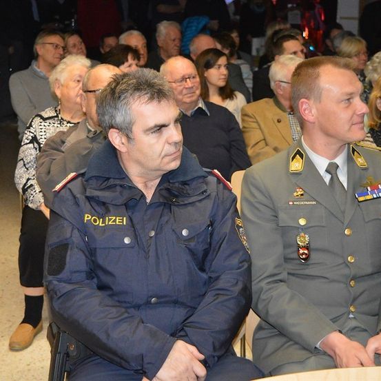 Zwei Männer in Uniform, einer in einer Polizeiuniform und der andere in einer Militäruniform, sitzen in einer Menschenmenge.
