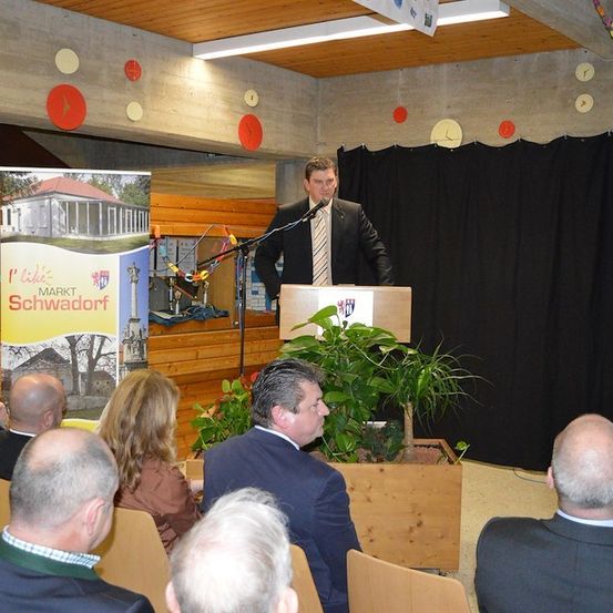 Ein Mann in Anzug und Krawatte spricht an einem Podium vor einem sitzenden Publikum. Ein Banner und Topfpflanzen sind hinter ihm.