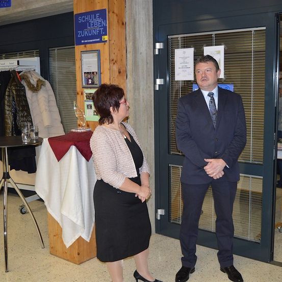 Ein Mann und eine Frau stehen vor einer Tür mit einem Schild, das 'SCHULE LEBEN ZUKUNFT' besagt. Die Frau trägt ein schwarzes Kleid und eine Brille, während der Mann einen Anzug und eine Krawatte trägt. Hinter ihnen steht ein Tisch mit einem Glas und einer Jacke darauf.