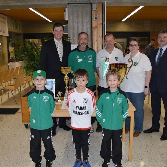 Eine Gruppe von Erwachsenen und Kindern steht zusammen und hält Trophäen in einer Halle mit Holzmöbeln und einer Fahne an der Wand.