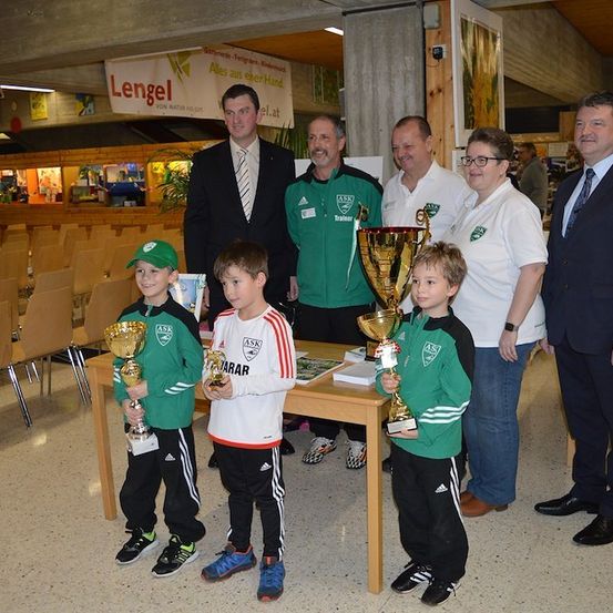 Eine Gruppe junger Jungen mit grünen Trikots und Mützen steht mit ihren Trophäen vor einem Tisch. Hinter ihnen stehen mehrere Erwachsene in formeller Kleidung. Die Umgebung scheint ein Sportereignis oder eine Preisverleihung zu sein.