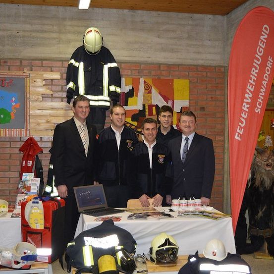 Fünf Männer in Anzügen stehen hinter einem Tisch mit Helmen und einem Banner mit der Aufschrift 'Feuerwehrjugend'. Ein Laptop und andere Gegenstände sind auf dem Tisch. Eine Feuerwehruniform hängt an der Wand.