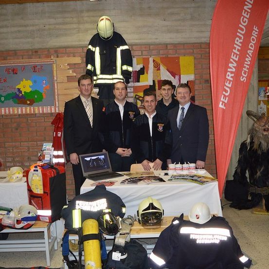 Fünf Männer in Anzügen stehen hinter einem Tisch mit Laptop und diverser Feuerwehrausrüstung. Ein Banner mit der Aufschrift 'Feuerwehrjugend'.
