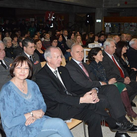 Eine Gruppe formell gekleideter Personen, darunter Männer und Frauen, sitzt in einem großen Auditorium. Einige tragen Brillen, Krawatten und Militäruniformen. Eine Projektionsleinwand ist vorne sichtbar.