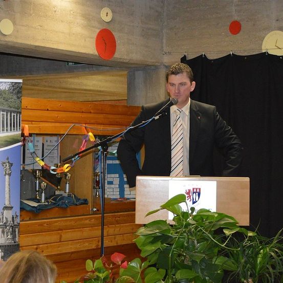 Ein Mann in Anzug und Krawatte steht an einem Podium mit Mikrofon und spricht bei einer Veranstaltung. Hinter ihm befindet sich eine Holzbühne mit Trophäen und einem Banner.