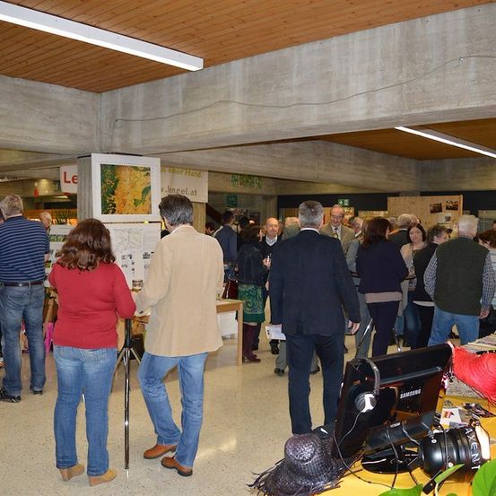 Eine Gruppe von Menschen ist in einem Raum versammelt, wahrscheinlich für eine Veranstaltung. Einige stehen, während andere herumlaufen. Eine Frau in einem roten Shirt schaut sich ein Poster an der Wand an. Ein Mann in einem Anzug geht auf den Schreibtisch mit einem Monitor zu. Es gibt auch einige Pflanzen und Hüte auf dem Schreibtisch.