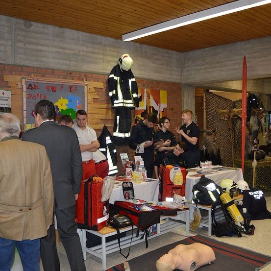 Eine Gruppe von Menschen versammelt sich in einem Raum mit verschiedenen ausgestellten Gegenständen, darunter ein Dummy, eine Feuerwehrmann-Uniform und eine rote Tasche. Einige Leute betrachten die Gegenstände, während andere sprechen.