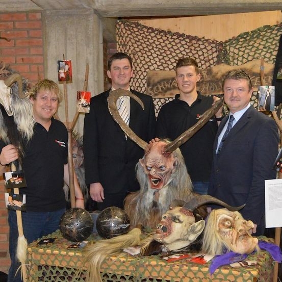 Vier Männer stehen hinter einem Tisch mit verschiedenen Masken und Requisiten, zwei von ihnen halten Hörner und Masken, einer hält einen Stock und einer hält ein Papier.
