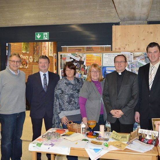 Fünf Personen, die formell gekleidet sind, stehen hinter einem Tisch mit verschiedenen Gegenständen, darunter ein Korb, eine Kerze und ein Becher. Sie scheinen für ein Foto zu posieren.