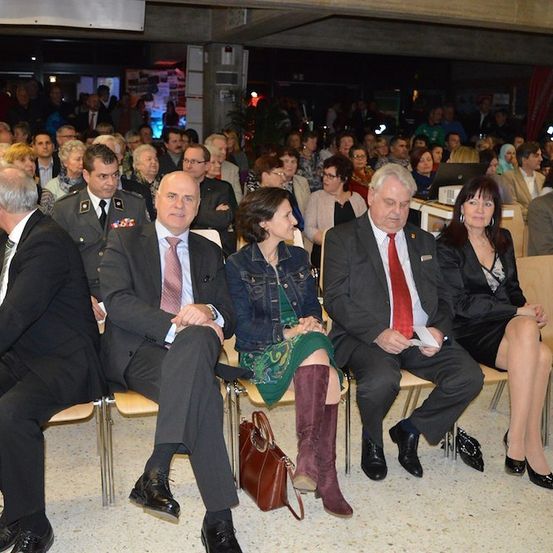 Eine Gruppe von Menschen in formeller Kleidung sitzt in Stühlen in einem Konferenzsaal, einige schauen auf Papiere. Eine Frau in einem grünen Kleid und hohen Stiefeln schaut nach vorne.