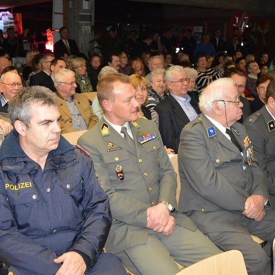 Eine Gruppe von Männern in Uniformen, einschließlich eines Polizeibeamten, sitzt unter einer Menge von Menschen in einem Saal.