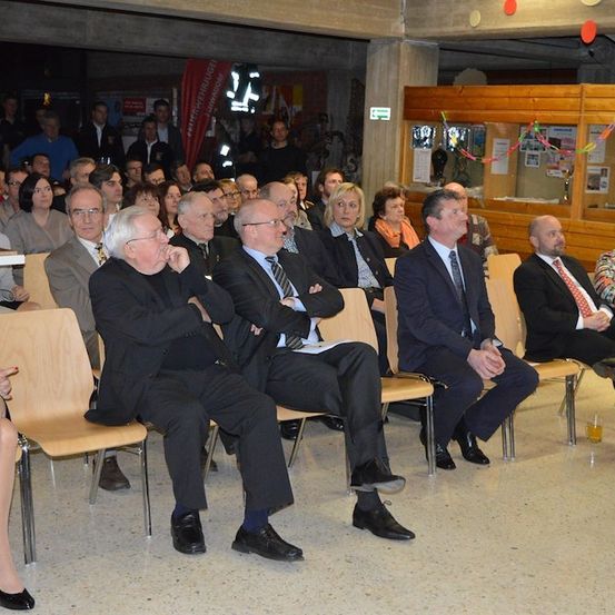 Eine Gruppe formell gekleideter Personen sitzt in einem großen Raum, möglicherweise bei einer formellen Veranstaltung. Einige schauen nach vorne, andere zur Seite. Im Hintergrund stehen Personen.
